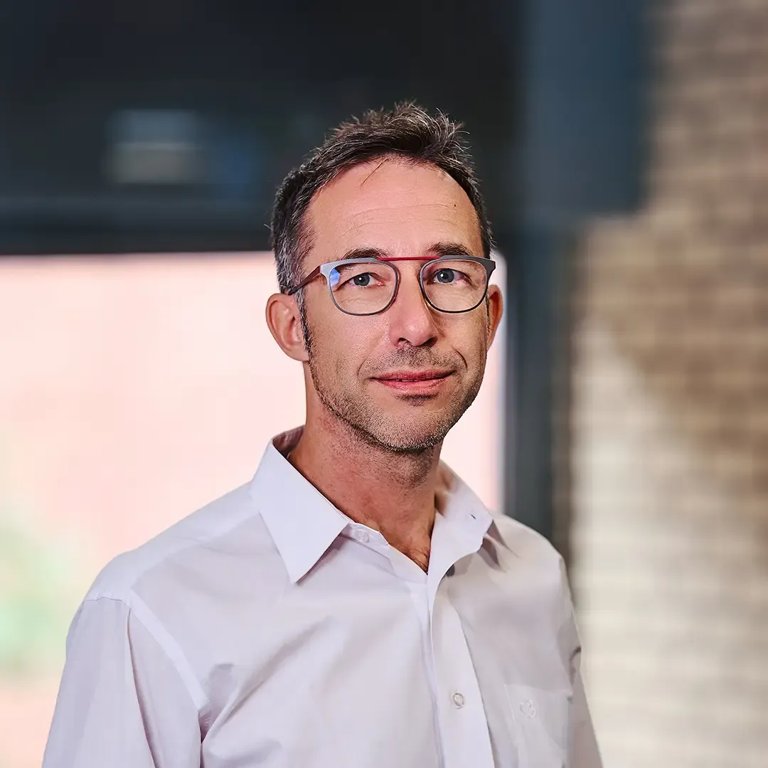Man with glasses in a white shirt stands indoors with a blurred background of a window and brick wall.