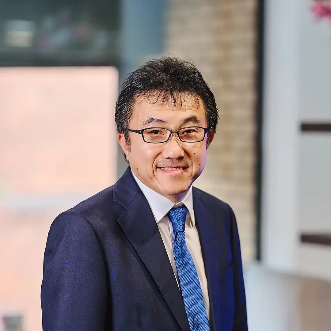 Smiling man in a suit and blue tie, standing in a modern office with a brick wall and window in the background.