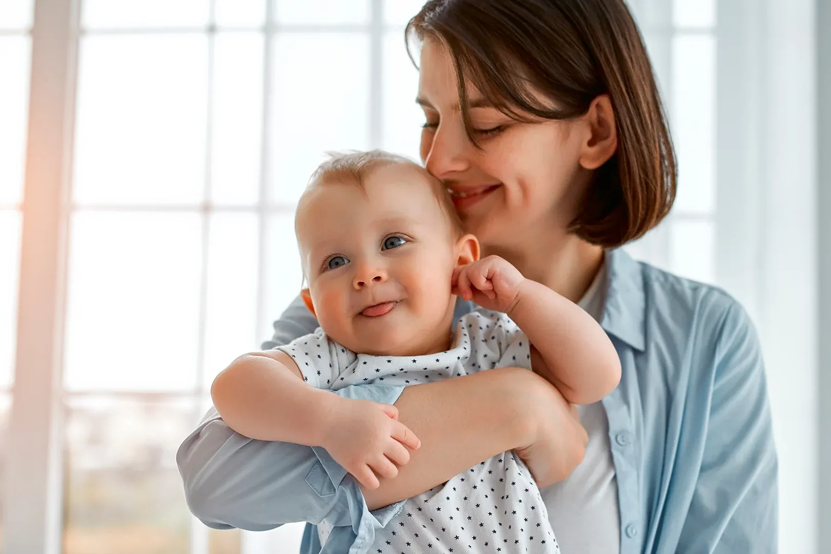 woman smiling toward her baby who is stood upright holding mothers arm