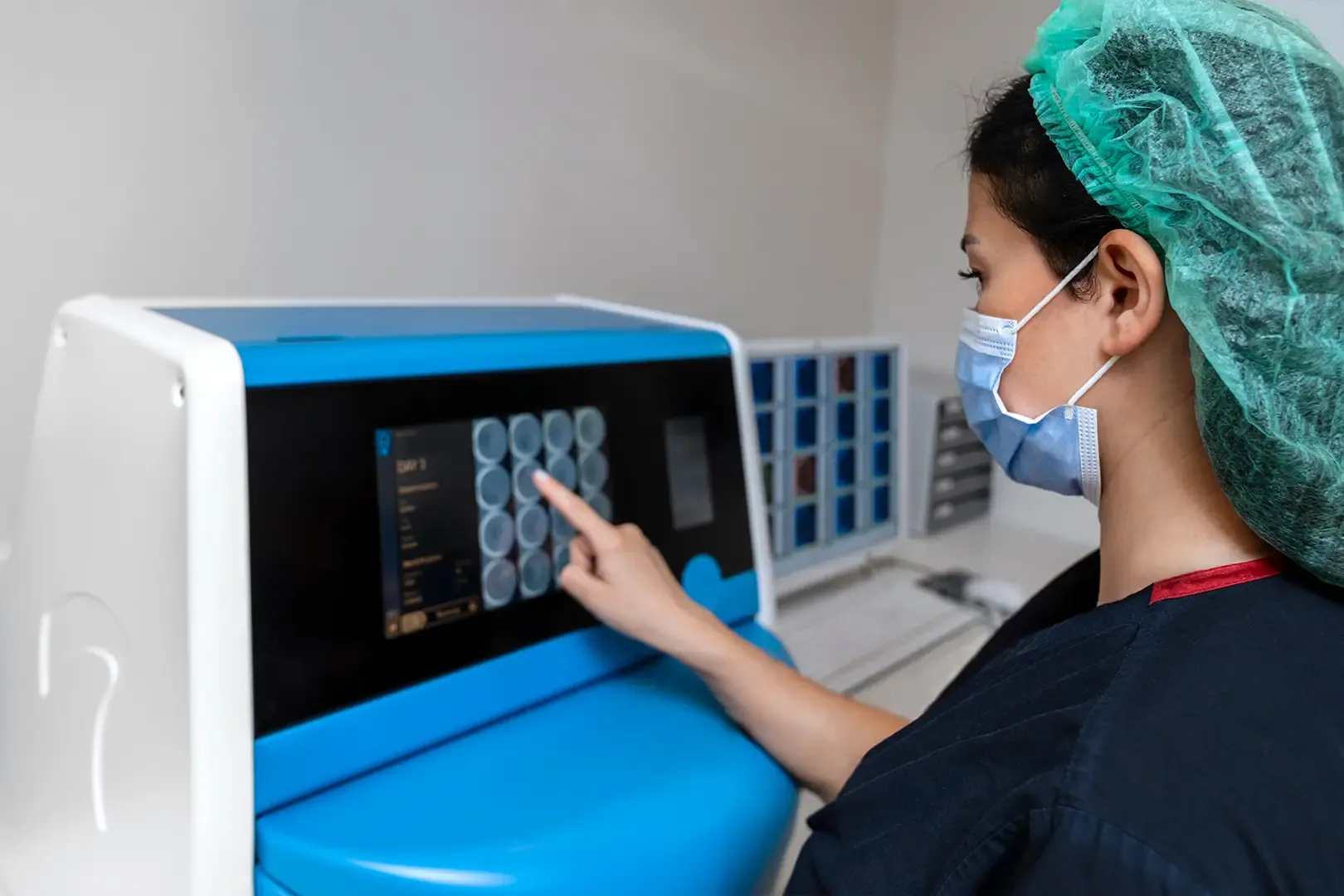 A healthcare worker in scrubs and a hairnet uses a touchscreen on a medical device in a clinical setting.