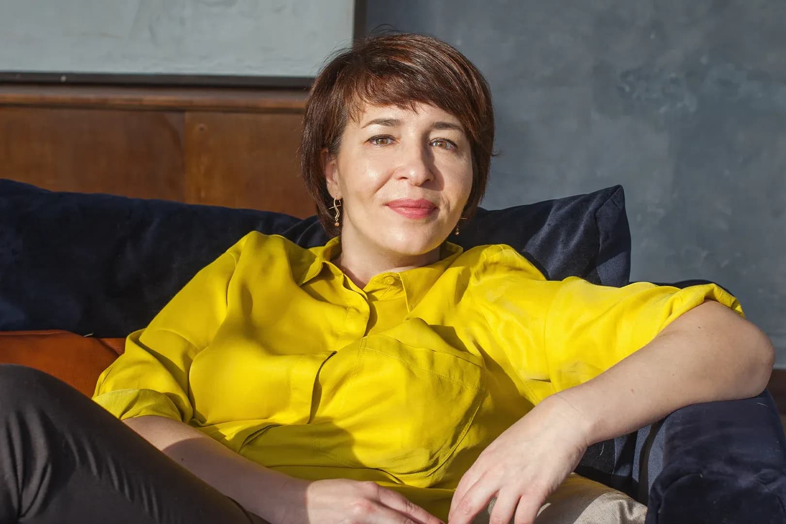 Woman in a bright yellow blouse sitting on a couch, smiling, with a relaxed pose in a cozy room with dark cushions and wooden accents.