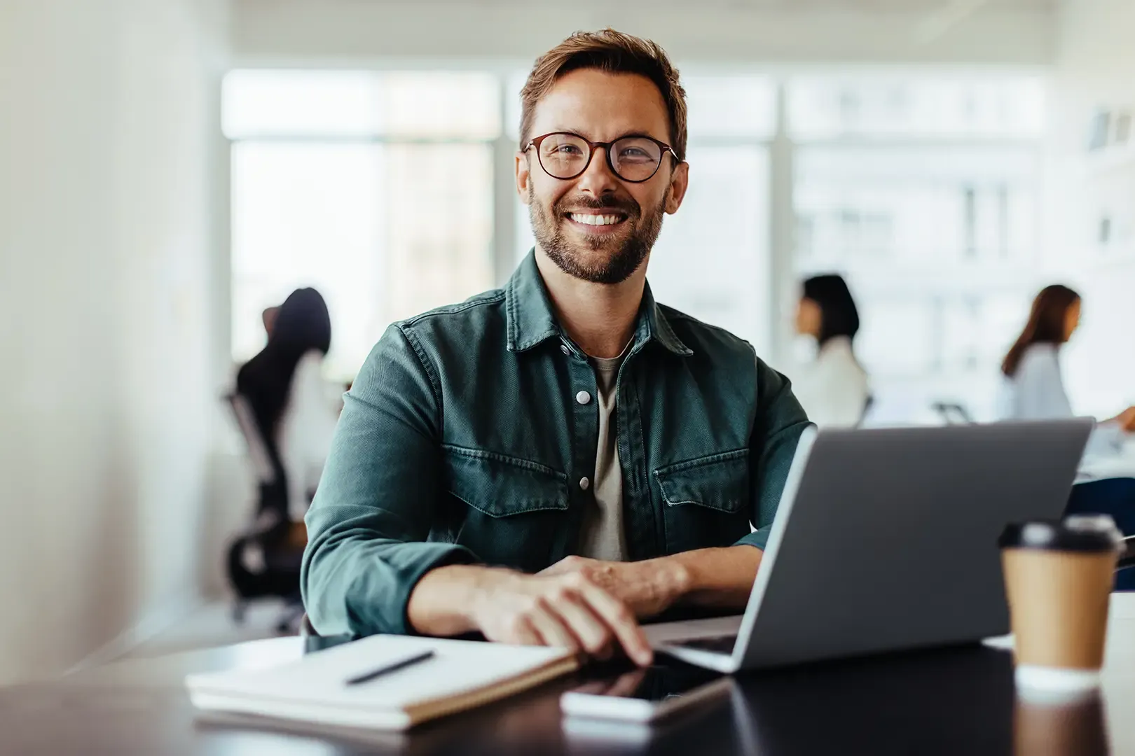 man with glasses sat at desk with laptop looks up towards camera and smiles