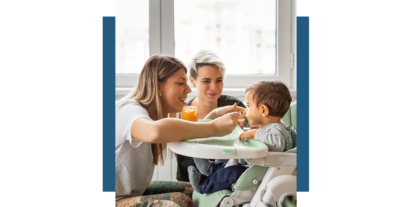 Female couple smiling and feeding their toddler in a high chair